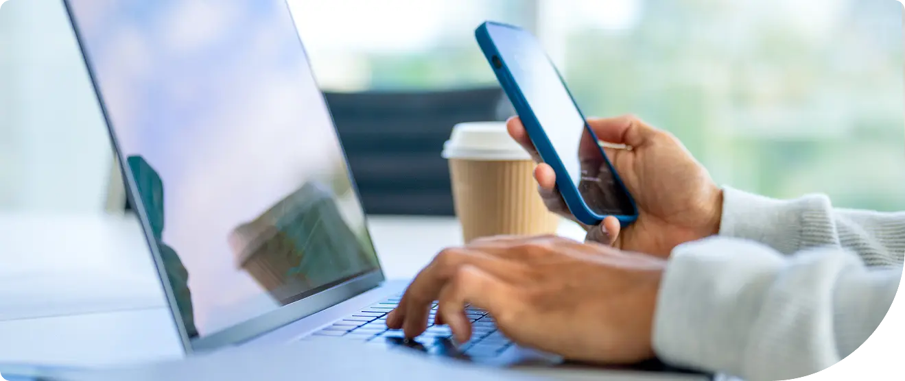 Close-up of a person&rsquo;s hands using a laptop while holding a smartphone, with a coffee cup on the desk in the background.