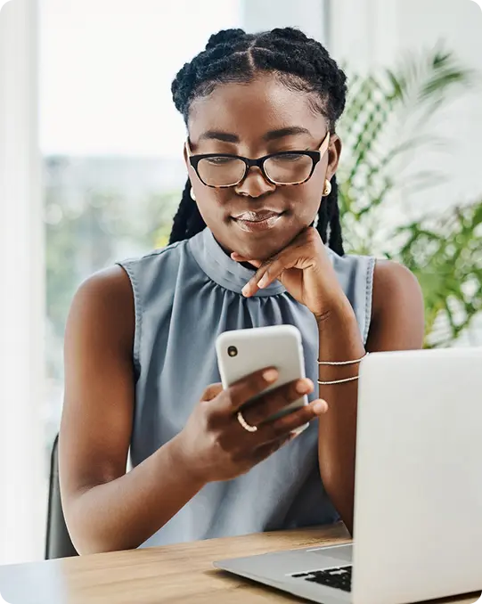 A woman wearing glasses sits at a desk with a laptop open in front of her, smiling while holding and looking at a smartphone.