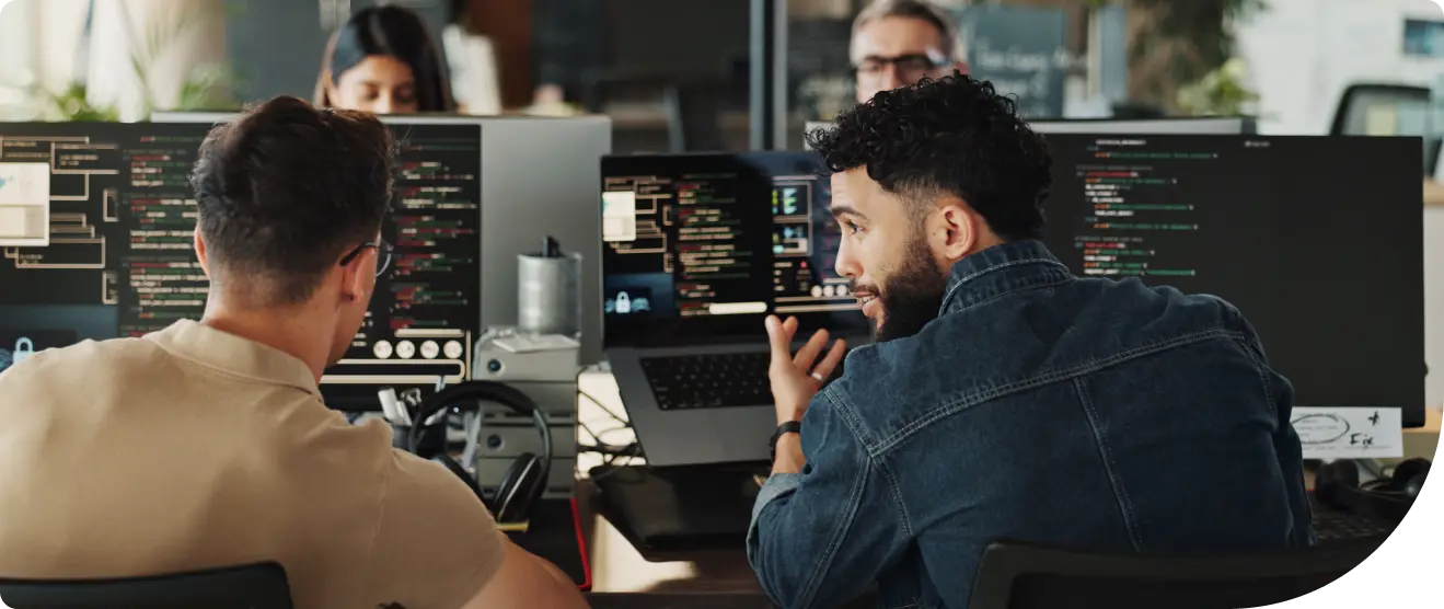 Two people review and discuss code on dual monitors, one typing and one pointing at the screen.