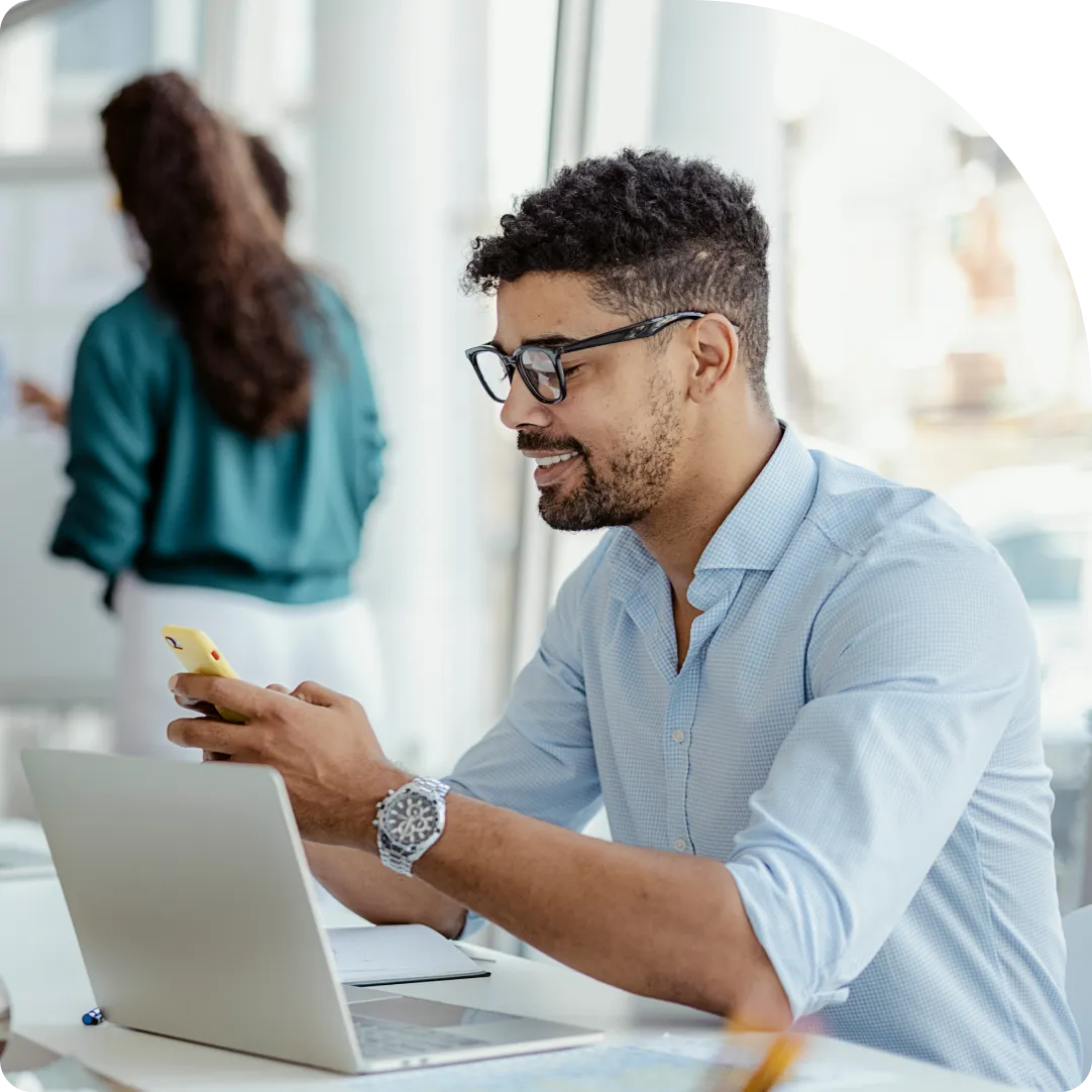 Smiling professional with glasses using Duo on a mobile device with their laptop on their desk.