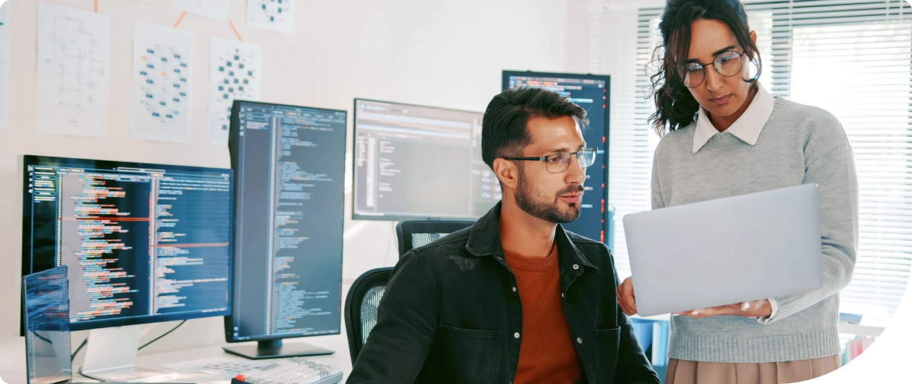 Two colleagues work together in a tech office, one seated at a desk with multiple monitors while the other holds a laptop