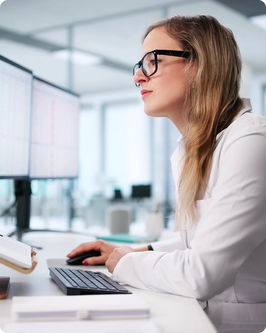 A woman wearing glasses looks at computer monitors in a bright, modern office