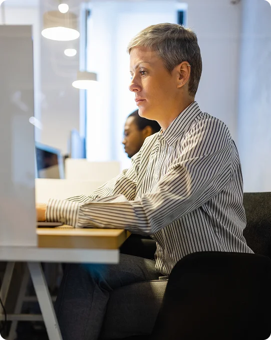 Woman with short gray hair working at a computer in a bright office environment with modern lighting and other employees nearby