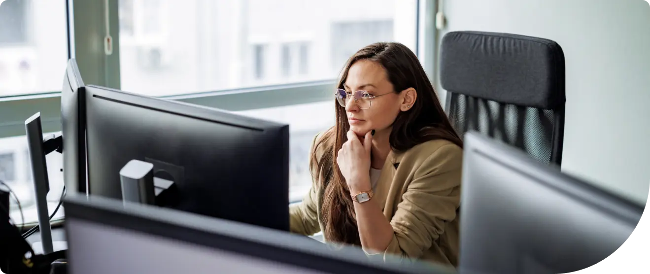 Woman sitting at a desk in an office, focused on a computer monitor with her hand resting on her chin in thought.