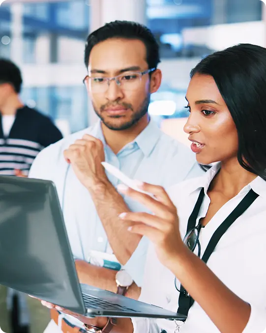 Two colleagues in an office review information on a laptop, while two others have a discussion in the background near workstations.