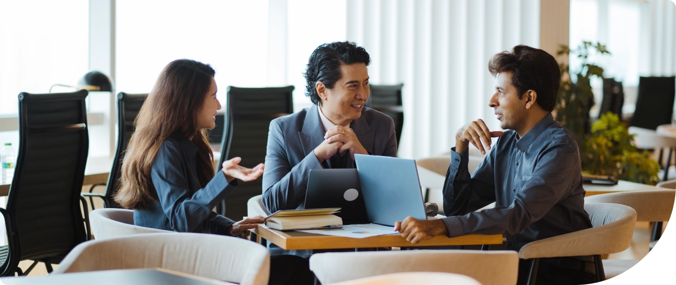 Three professionals sit together at a table in a modern office, engaged in a meeting with laptops and notebooks open