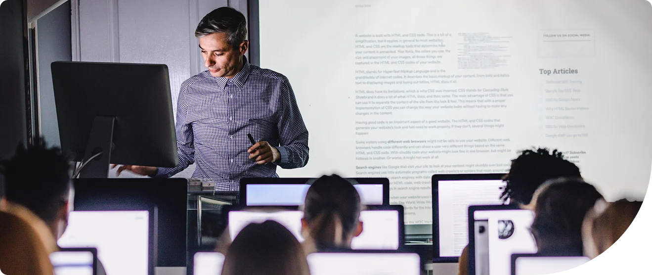 Man leading a lecture style meeting with notes on a screen at the front of the room and an audience on their laptops