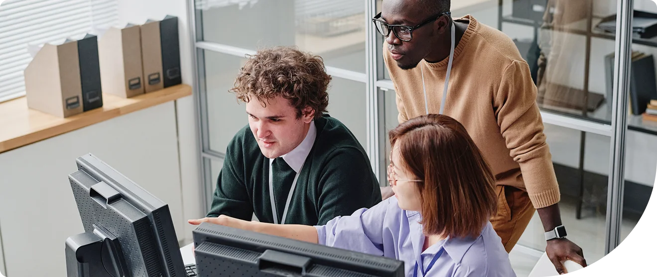 Woman pointing at something on her desk with a colleague beside and behind her look at what she&rsquo;s pointing at, outside of the image frame