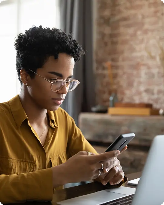 A woman wearing glasses sits at a desk, using a smartphone and a laptop open in front of her