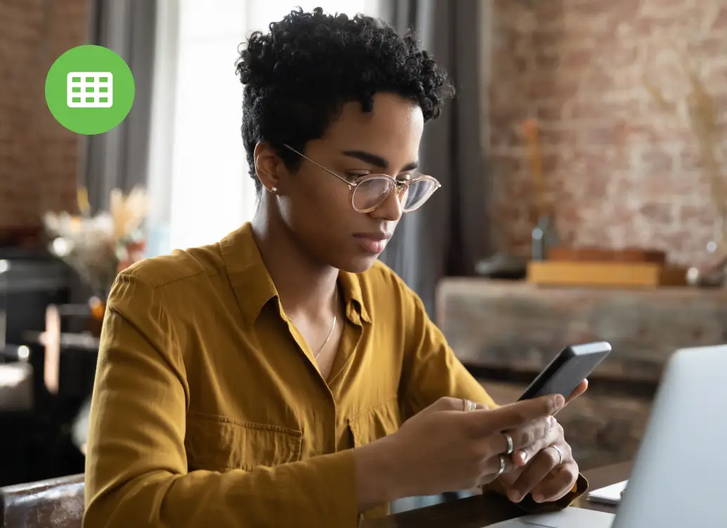 A woman wearing glasses sits at a desk, using a smartphone and a laptop open in front of her