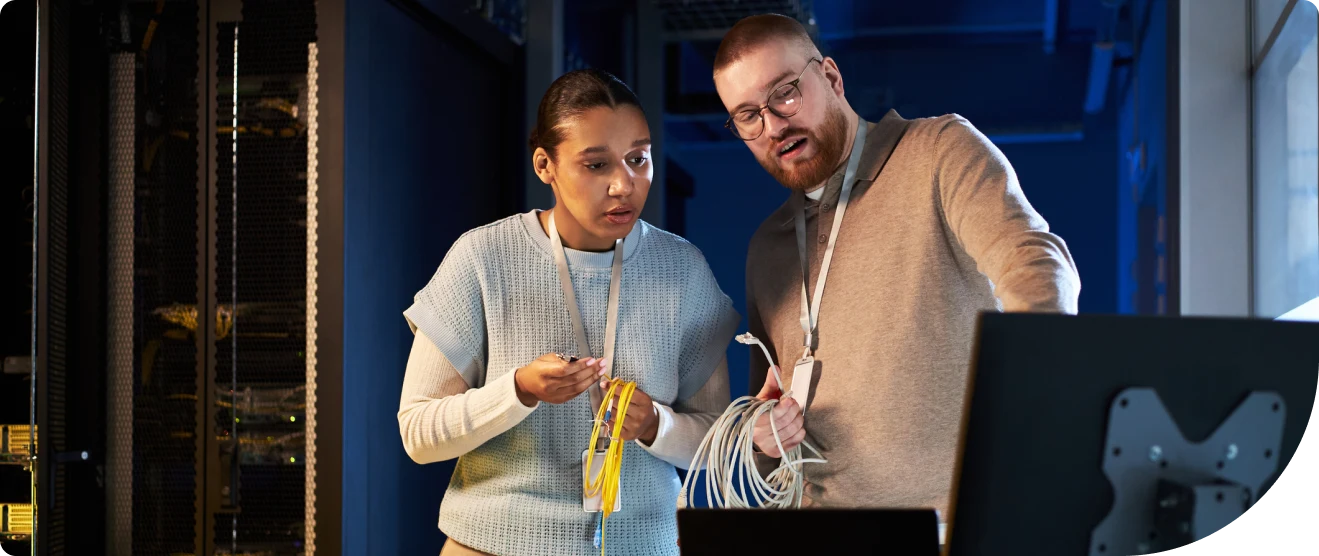 Two IT professionals inspect network cables and a computer monitor in a server room