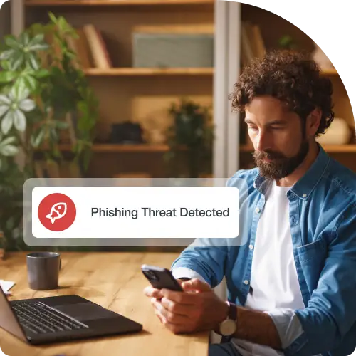 A man working on a laptop at a desk in his home office is looking at his mobile phone. A digital message box on the left side of the image displays a Phishing icon and the text Phishing Threat Detected.