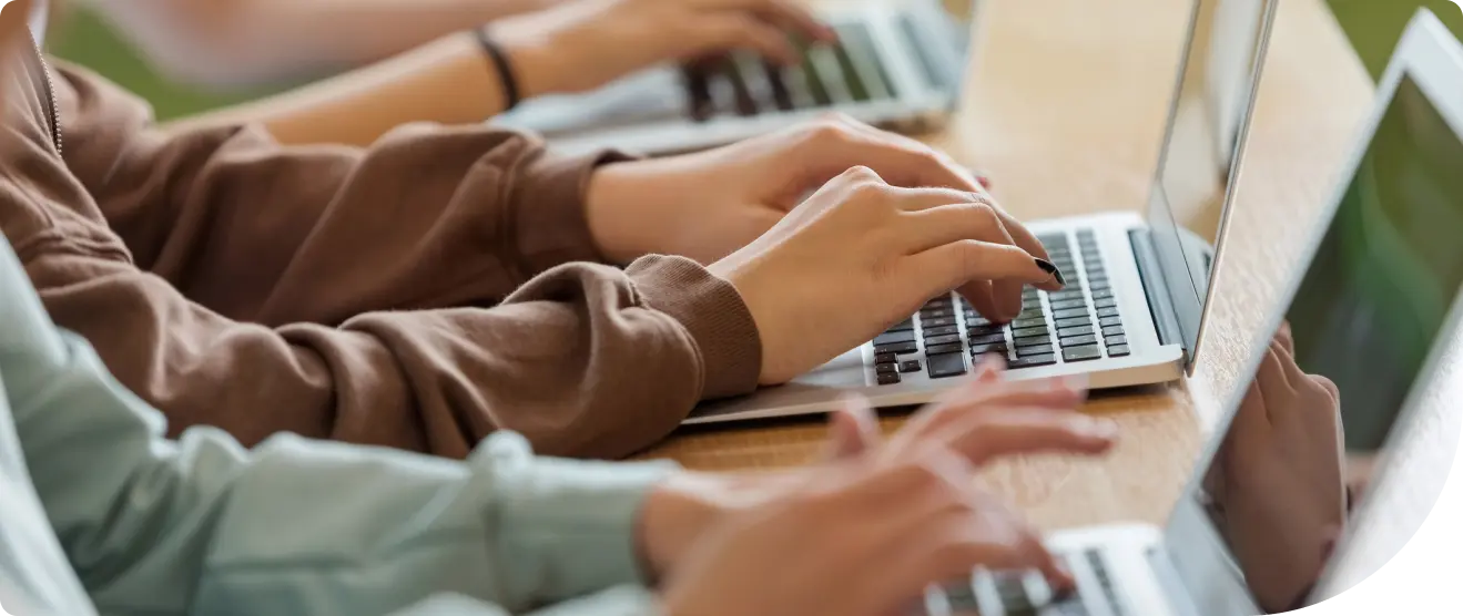 Close-up of several people typing on laptops at a wooden desk, with hands and keyboards in focus during group computer work.