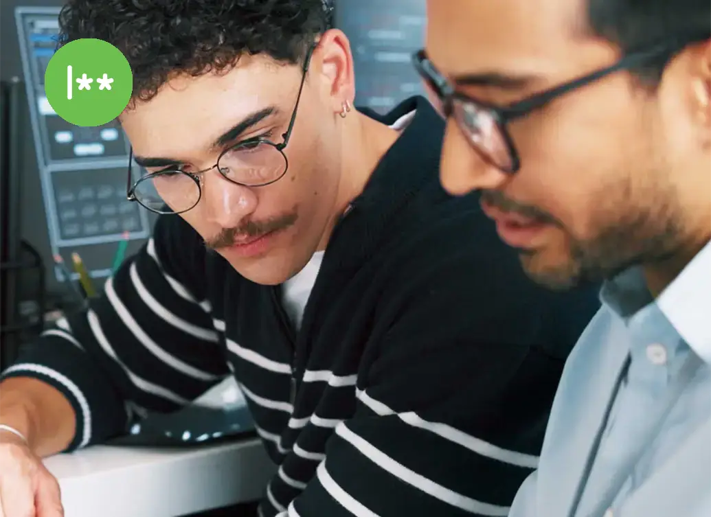 Two colleagues review information on a tablet in front of multiple computer monitors displaying code and technical data.