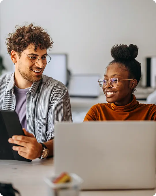 Two coworkers smiling and talking at their desks, one holding a tablet and the other typing on a laptop in a modern office setting.