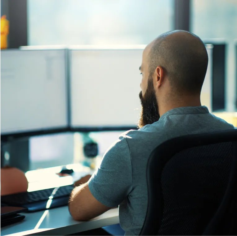 Person at a desk with multiple monitors in a sunlit room.