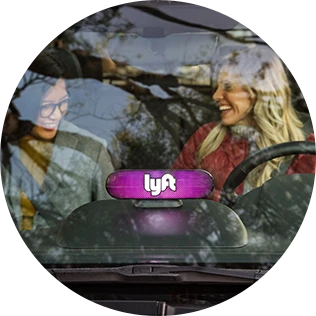 A happy Lyft driver welcomes her customer, a bright pink Lyft sign is mounted on the car's dashboard