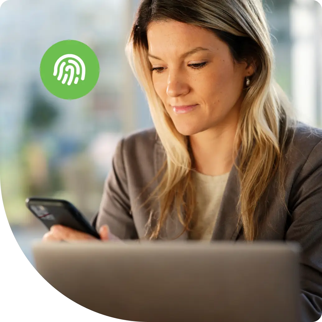 A professional woman authenticating on a smartphone while working at a laptop in a bright office