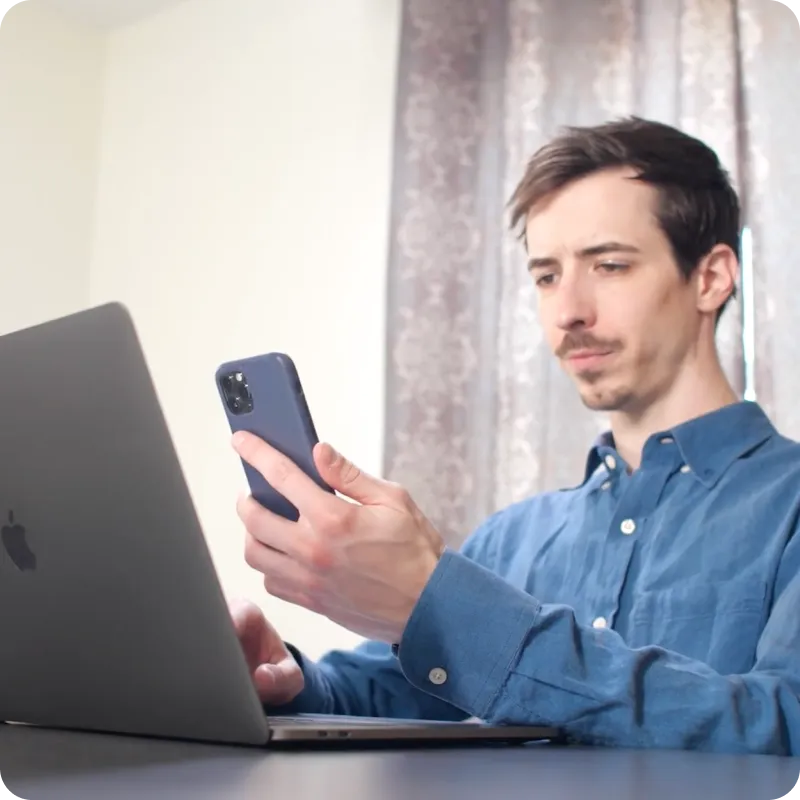 A person in a blue shirt is sitting at a desk using a laptop and holding a smartphone.