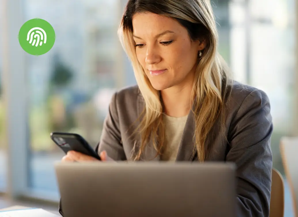A professional woman authenticating on a smartphone while working at a laptop in a bright office