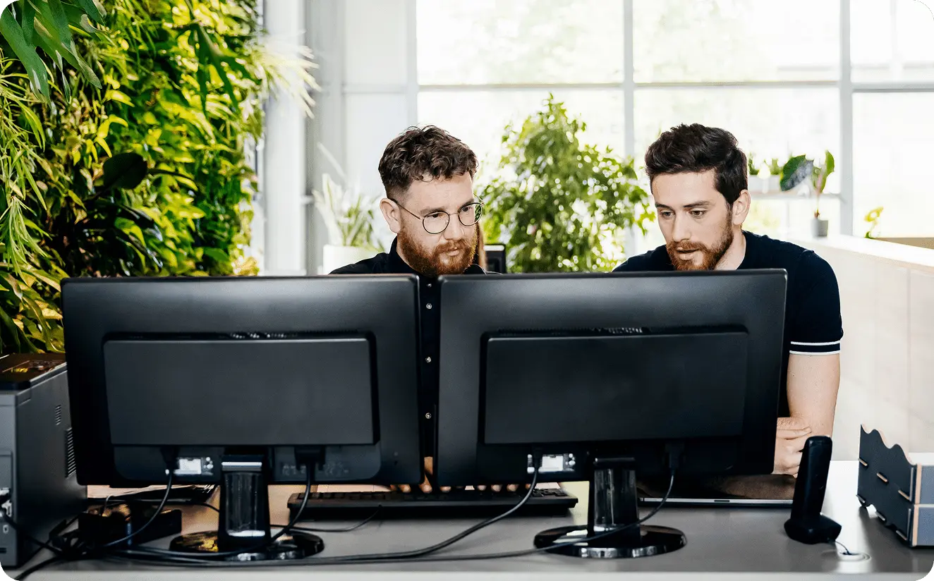 Two men working together at a desk with dual monitors in a modern office featuring a green plant wall.
