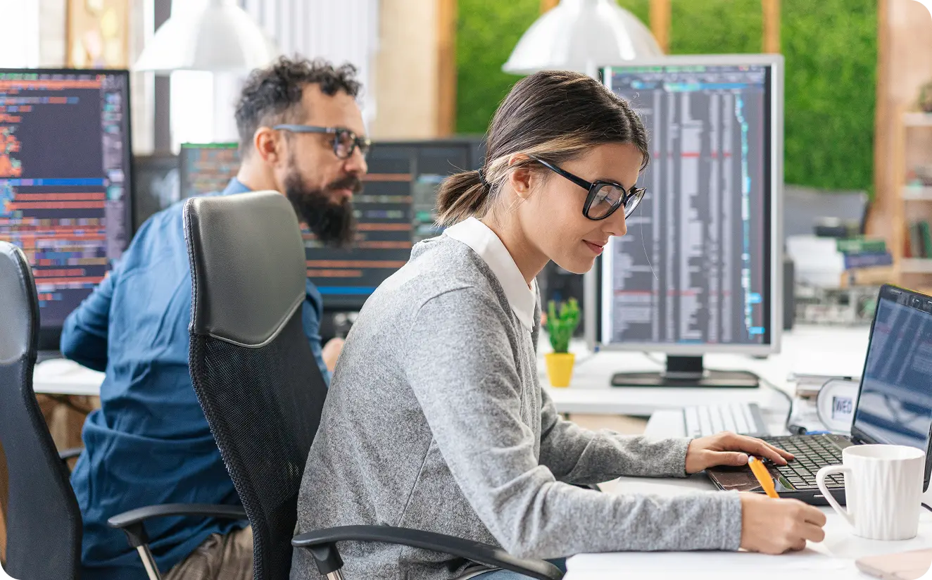 A woman writing notes while working on a laptop, with a man coding on desktop computers in the background in a modern office.